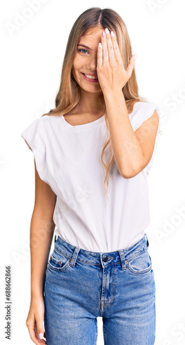 Beautiful caucasian woman with blonde hair wearing casual white tshirt covering one eye with hand, confident smile on face and surprise emotion.