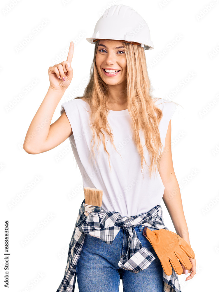 Beautiful caucasian woman with blonde hair wearing hardhat and painter clothes pointing finger up with successful idea. exited and happy. number one. Beautiful caucasian woman with blonde hair wearing hardhat and painter clothes pointing finger up with successful idea. exited and happy. number one.