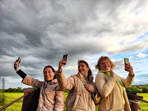 Three funny tourist girls on the old bridge take selfies against the background of an alarming dramatic sky with thunderclouds. Middle-aged women having fun outdoors