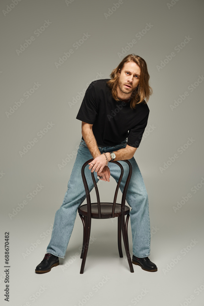 full length of bearded good looking man with long hair in jeans and t-shirt sitting on chair on grey