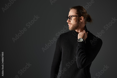 Fototapeta Naklejka Na Ścianę i Meble -  handsome man in eyeglasses pulling high collar on turtleneck sweater while posing on grey background