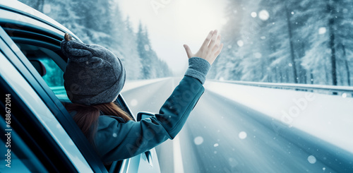 Happy woman in car waving while driving through winter forest. Woman driver feels wind through her hands while driving in winter landscape. Winter vacation and freedom concept