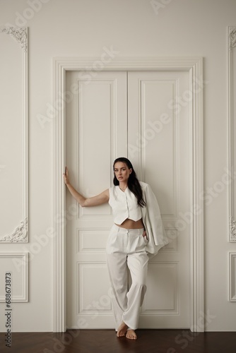 A young brunette woman wearing a white suit, posing in a photo studio with a bright interior.