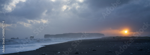 Reynisfjara black sand beach at sunset. Moddy skies and golden sun. A couple (small) walking along beach. South Coast of iceland. Panorama image.