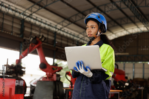 Portrait of female mechanical engineer worker in yellow hard hat and safety uniform using laptop standing at manufacturing area of industrial factory