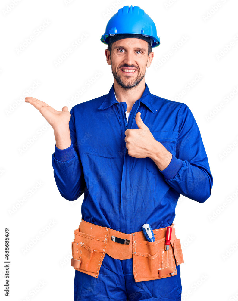 Young handsome man wearing worker uniform and hardhat showing palm hand and doing ok gesture with thumbs up, smiling happy and cheerful