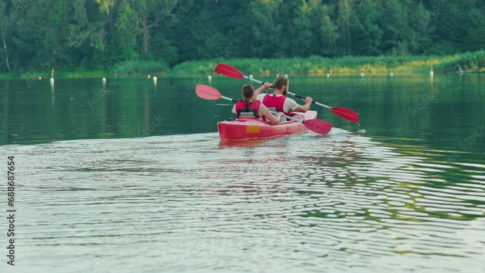 Backside view of couple practicing rowing together on lake. Man and woman with red vests sitting in boat and rowing with oars on water. Entertainment outdoors. Canoeing workout. Activity concept. Backside view of couple practicing rowing together on lake. Man and woman with red vests sitting in boat and rowing with oars on water. Entertainment outdoors. Canoeing workout. Activity concept.