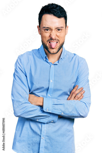 Hispanic man with beard with arms crossed gesture sticking tongue out happy with funny expression.