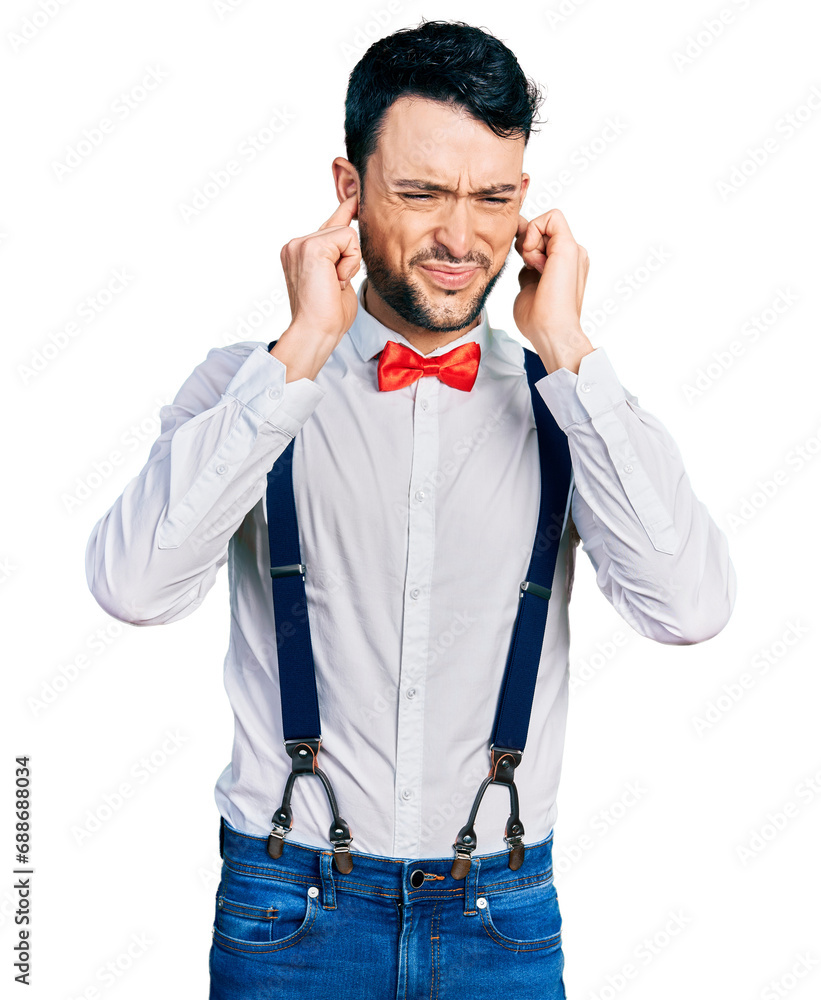 Hispanic man with beard wearing hipster look with bow tie and suspenders covering ears with fingers with annoyed expression for the noise of loud music. deaf concept. Hispanic man with beard wearing hipster look with bow tie and suspenders covering ears with fingers with annoyed expression for the noise of loud music. deaf concept.