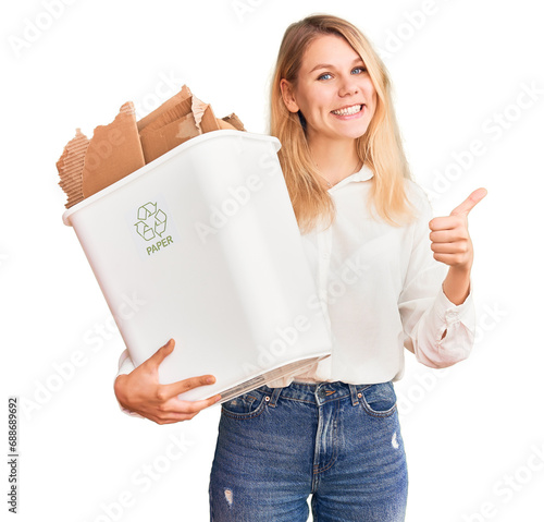 Young beautiful blonde woman holding recycle paper bin smiling happy and positive, thumb up doing excellent and approval sign photo