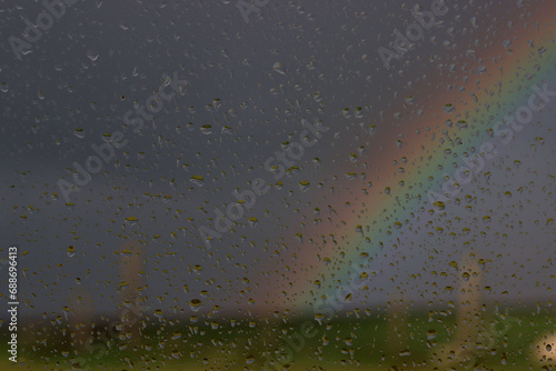 Focus on  rain drops on a window. In the distance intentionally out of focus, very dark storm clouds and a rainbow. 