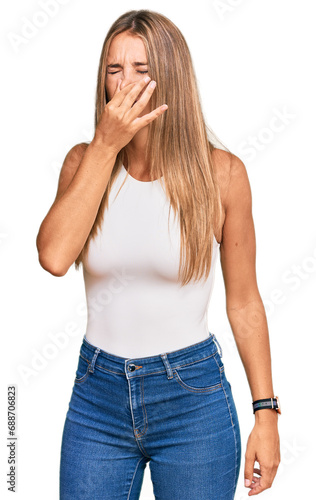 Young blonde woman wearing casual style with sleeveless shirt smelling something stinky and disgusting, intolerable smell, holding breath with fingers on nose. bad smell