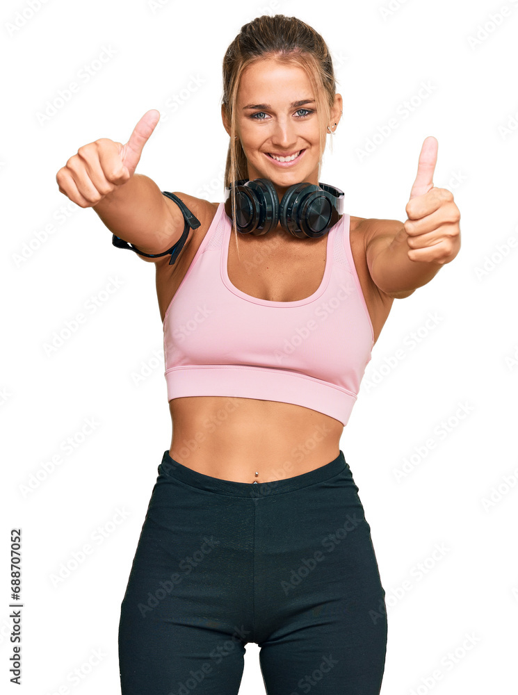 Young blonde woman wearing gym clothes and using headphones approving doing positive gesture with hand, thumbs up smiling and happy for success. winner gesture. Young blonde woman wearing gym clothes and using headphones approving doing positive gesture with hand, thumbs up smiling and happy for success. winner gesture.