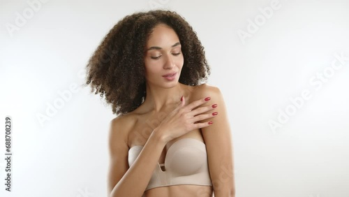 Curly-haired thin build sexy beauty wearing nude strapless bra running her fingers with red manicure on perfect body standing on white background. High quality 4k footage