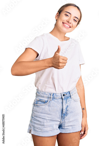 Young beautiful blonde woman wearing casual white tshirt doing happy thumbs up gesture with hand. approving expression looking at the camera showing success.