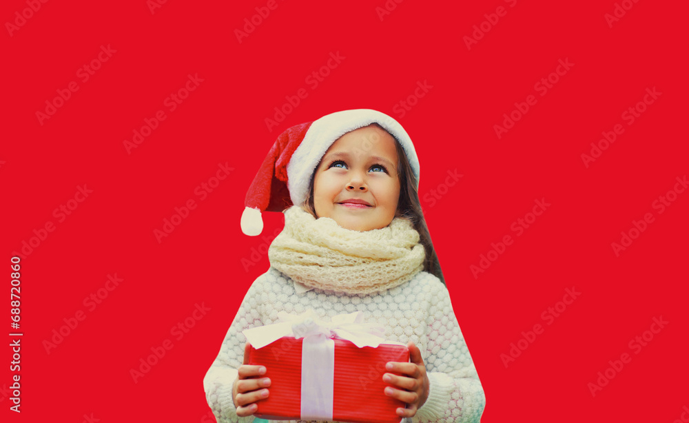 Fototapeta premium Christmas portrait of happy smiling little girl child in santa red hat with gift box and looks up isolated on studio background