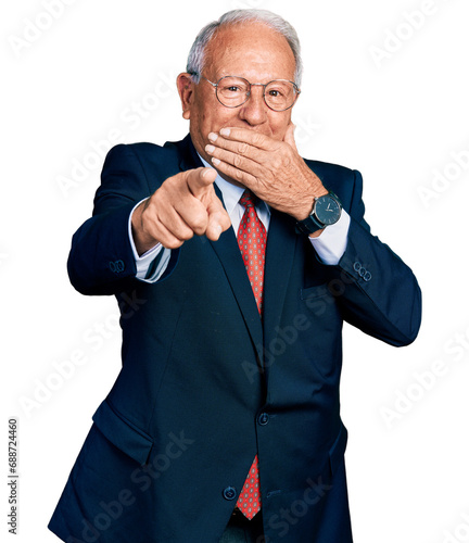 Senior man with grey hair wearing business suit and glasses laughing at you, pointing finger to the camera with hand over mouth, shame expression