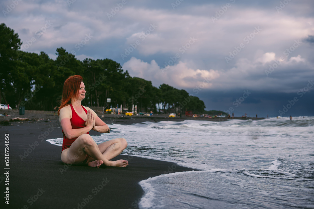 Happy woman tourist in red bodysuit Sitting in the lotus position, yoga on black sand, stormy waves on the sea. Danger for swimming. Rain clouds on the horizon. Copy space. Ureki besch, Georgia