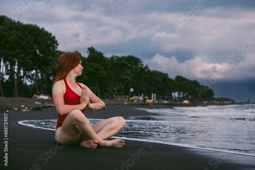 Happy woman tourist in red bodysuit Sitting in the lotus position, yoga on black sand, stormy waves on the sea. Danger for swimming. Rain clouds on the horizon. Copy space. Ureki besch, Georgia