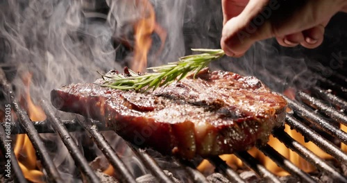 Ribeye steak sizzles on grill seasoned with rosemary. Tongues of flame break through grill grate. Aroma of juicy Ribeye steak wafts through air. Grill brings out best in every bite, close-up cinematic
