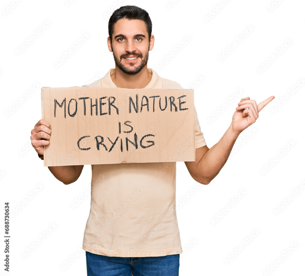 Young hispanic man holding mother nature is crying protest cardboard banner smiling happy pointing with hand and finger to the side Young hispanic man holding mother nature is crying protest cardboard banner smiling happy pointing with hand and finger to the side