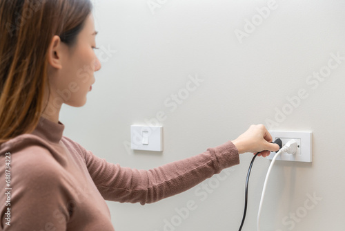 Fotografie Asian young woman plugged in, unplugged electricity cord cable, put on or remove electric plug cable in socket on wall home outlet for saving, control power electrical energy, eco environment concept