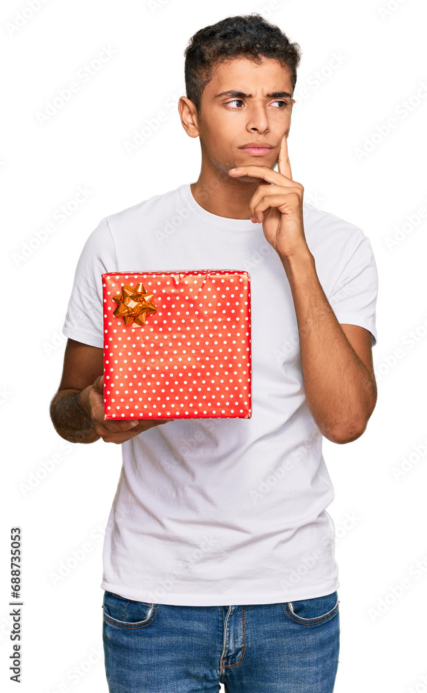 Young handsome african american man holding gift serious face thinking about question with hand on chin, thoughtful about confusing idea Young handsome african american man holding gift serious face thinking about question with hand on chin, thoughtful about confusing idea