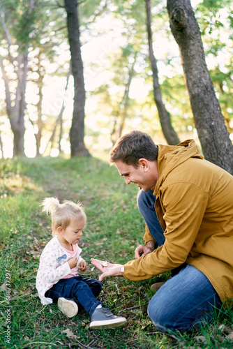Dad holds out a tiny mushroom in the palm to a little girl sitting in the forest on the grass © Nadtochiy