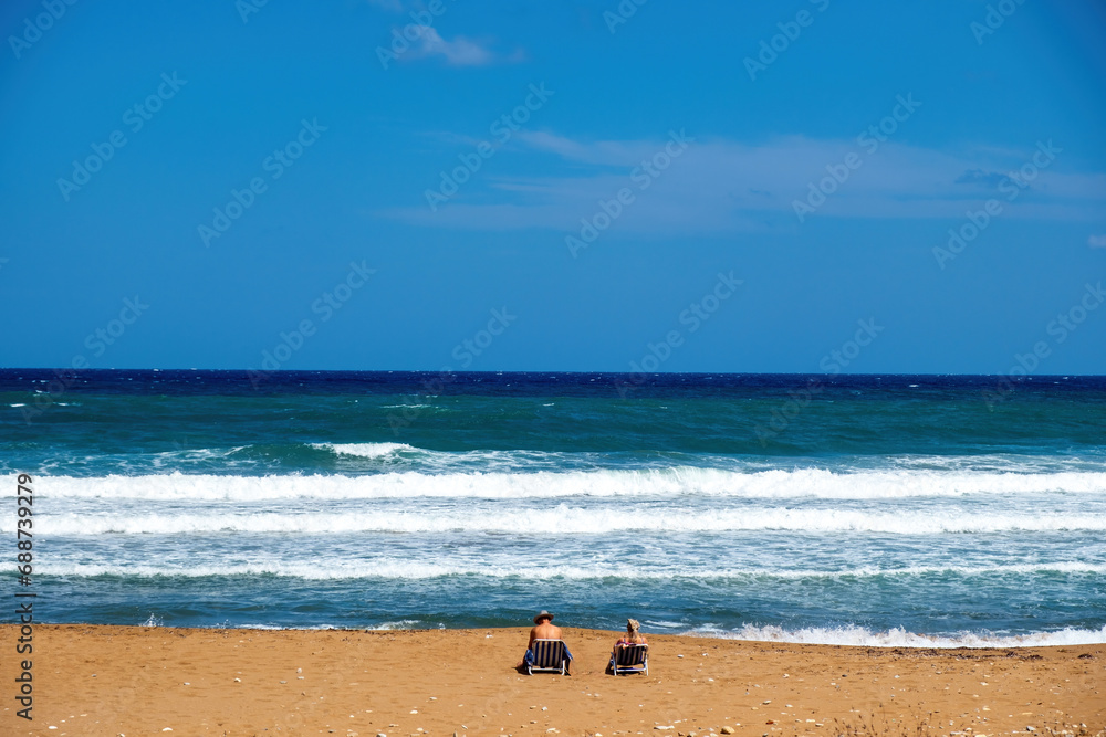 Greek sandy beach. Ripple sea water, splashing foam, blue sky background, couple relax at seaside.