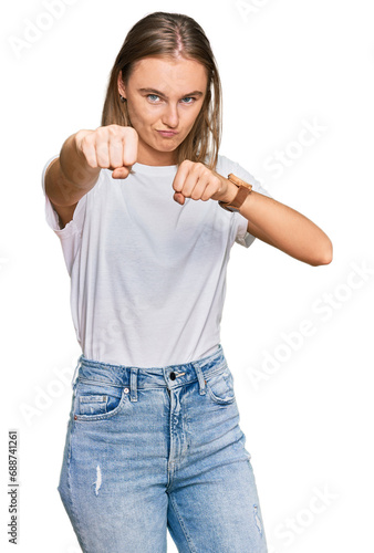 Beautiful young blonde woman wearing casual white t shirt punching fist to fight, aggressive and angry attack, threat and violence