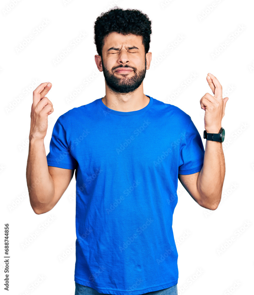 Fototapeta premium Young arab man with beard wearing casual blue t shirt gesturing finger crossed smiling with hope and eyes closed. luck and superstitious concept.