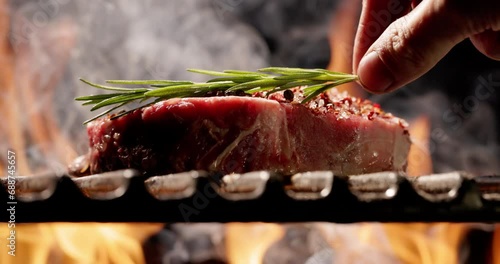 Close-up. Hand gently places sprig of fragrant rosemary onjuicy steak sprinkled with seasonings. Flickering fire of barbecue sets stage for unforgettable summer party. Flickering flames in background. photo