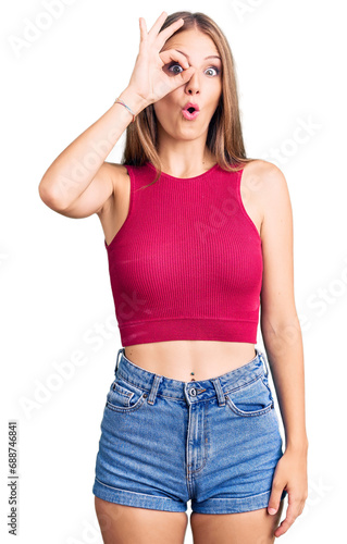 Young beautiful blonde woman wearing elegant summer shirt doing ok gesture shocked with surprised face, eye looking through fingers. unbelieving expression.