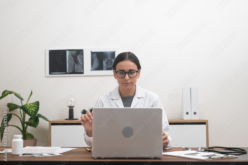 Young female doctor reviews examination of patient organs on laptop medical specialist works sitting at table with pictures and documents in modern clinic Young female doctor reviews examination of patient organs on laptop medical specialist works sitting at table with pictures and documents in modern clinic