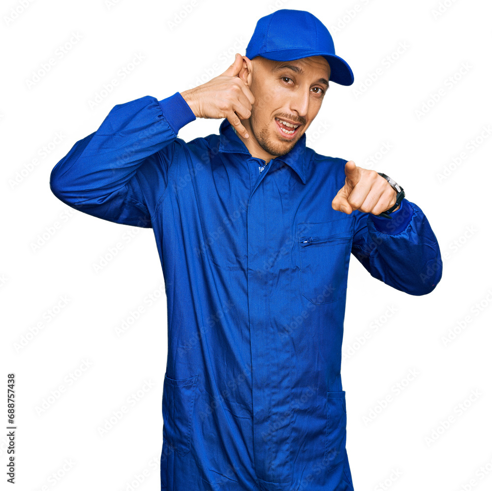 Bald man with beard wearing builder jumpsuit uniform smiling doing talking on the telephone gesture and pointing to you. call me.
