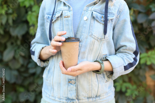 Woman in jeans jacket holding cardboard coffee cup against green plants wall background