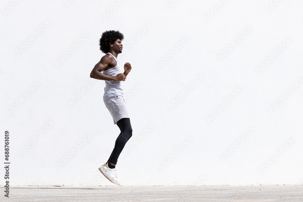 Side view of determined young active African American male athlete in sportswear jumping on street against white wall during fitness workout Side view of determined young active African American male athlete in sportswear jumping on street against white wall during fitness workout