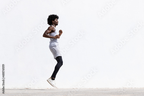 Side view of determined young active African American male athlete in sportswear jumping on street against white wall during fitness workout photo