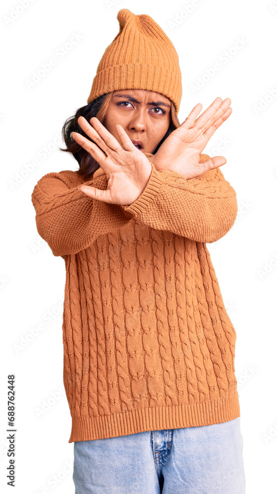 Young beautiful mixed race woman wearing wool sweater and winter hat rejection expression crossing arms doing negative sign, angry face Young beautiful mixed race woman wearing wool sweater and winter hat rejection expression crossing arms doing negative sign, angry face