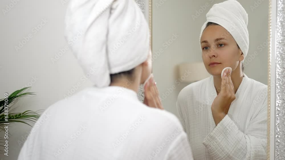 A woman, in her bathroom sanctuary, cares for her skin by cleansing with a cotton pad near the mirror, radiating beauty and self-care. A woman, in her bathroom sanctuary, cares for her skin by cleansing with a cotton pad near the mirror, radiating beauty and self-care.