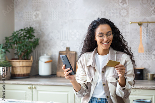 Young beautiful woman in kitchen with phone and bank credit debit card in hands, smiling Hispanic woman chooses gifts and products in online store, online shopping remotely from home.