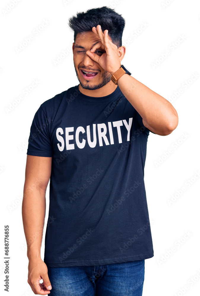 Handsome latin american young man wearing security t shirt doing ok gesture with hand smiling, eye looking through fingers with happy face. Handsome latin american young man wearing security t shirt doing ok gesture with hand smiling, eye looking through fingers with happy face.