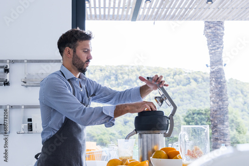 Side view of bearded man in apron standing near table with orange fruits squeezer in modern kitchen while preparing orange juice photo