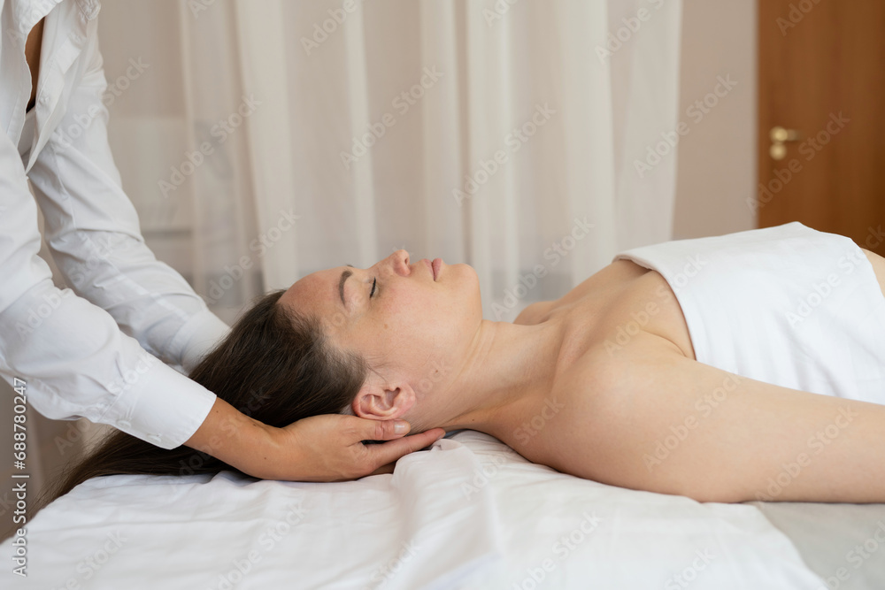 Side view of female customer wrapped in white towel with closed eyes lying on bed and receiving neck therapy massage from unrecognizable message therapist Side view of female customer wrapped in white towel with closed eyes lying on bed and receiving neck therapy massage from unrecognizable message therapist