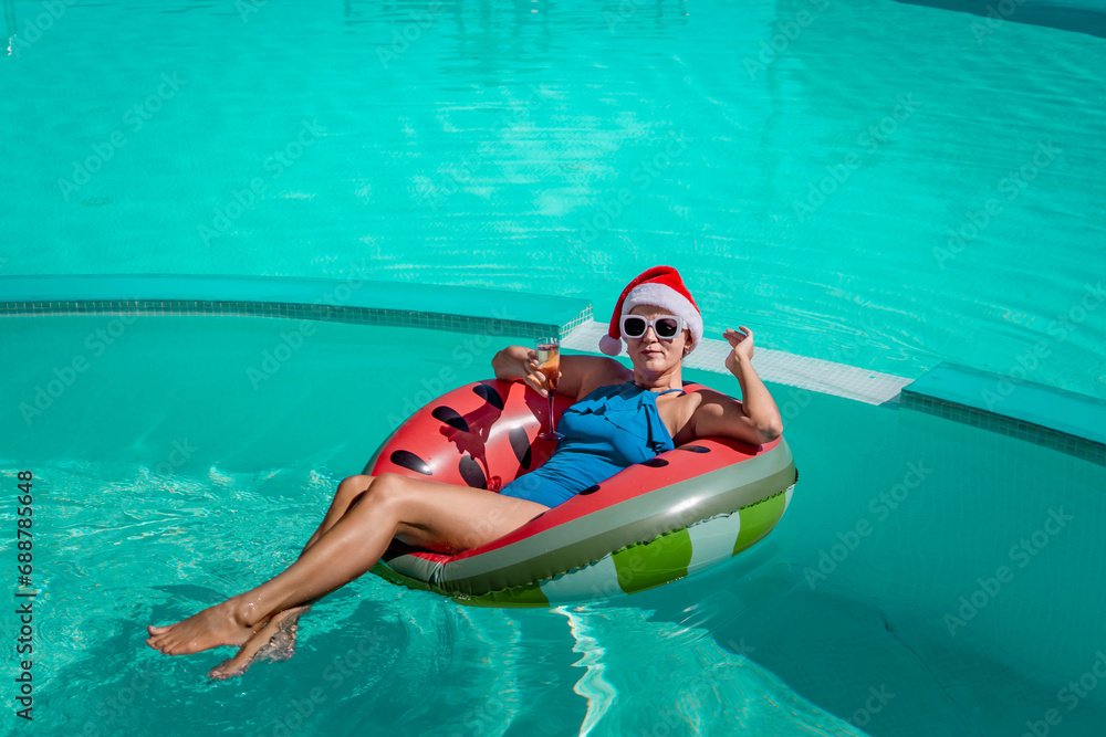 A happy woman in a blue bikini, a red and white Santa hat and sunglasses poses in the pool in an inflatable circle with a watermelon pattern, holding a glass of champagne in her hands. Christmas