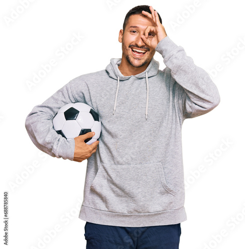 Handsome man with tattoos holding soccer ball smiling happy doing ok sign with hand on eye looking through fingers