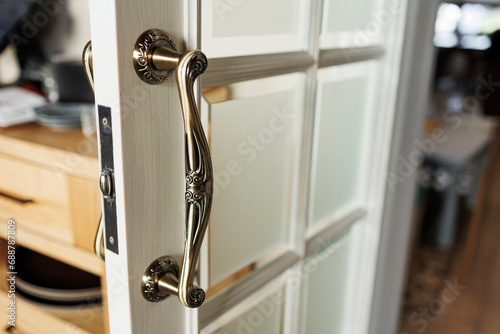 Close up of stylish stainless door knob or handle with metallic carvings on open white wooden modern interior door with glass. Concept of trends interior details.