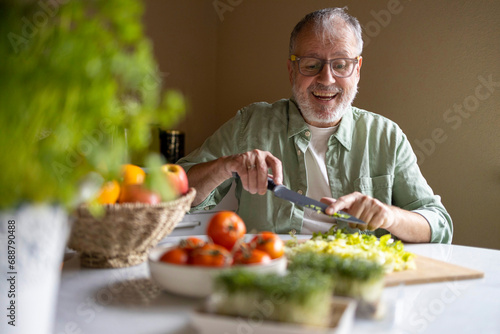 Cheerful senior man in glasses eyewear cutting lettuce in pieces with knife on wooden chopping board while preparing salad at home photo