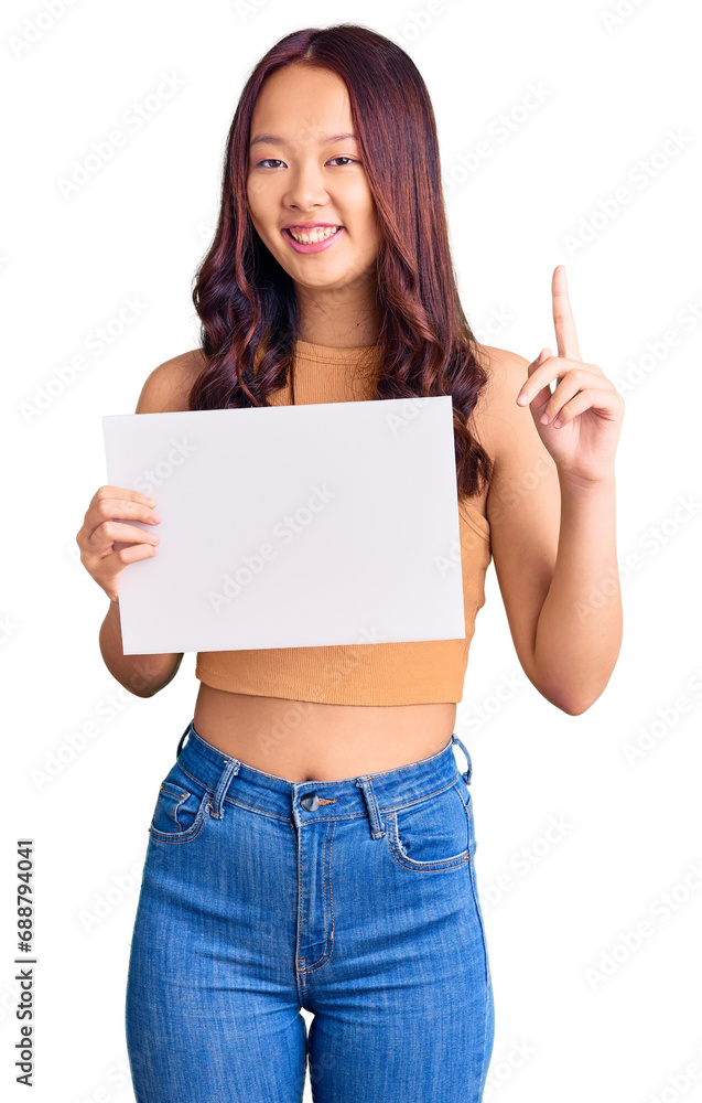 Young beautiful chinese girl holding paper banner with blank space surprised with an idea or question pointing finger with happy face, number one Young beautiful chinese girl holding paper banner with blank space surprised with an idea or question pointing finger with happy face, number one