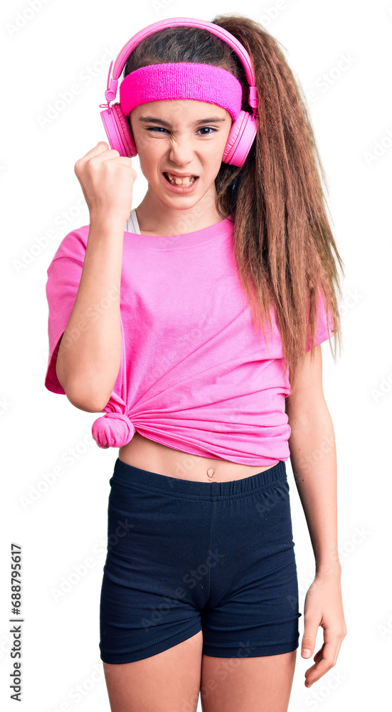 Cute hispanic child girl wearing gym clothes and using headphones angry and mad raising fist frustrated and furious while shouting with anger. rage and aggressive concept. Cute hispanic child girl wearing gym clothes and using headphones angry and mad raising fist frustrated and furious while shouting with anger. rage and aggressive concept.
